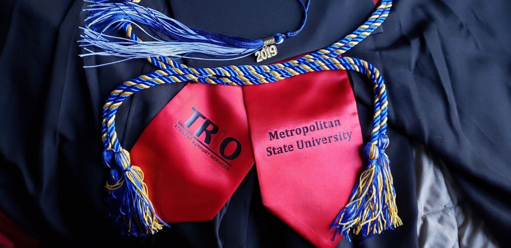 A red gratitude sash, blue and gold honors cord, blue and white 2019 tassel on a black graduation goan
